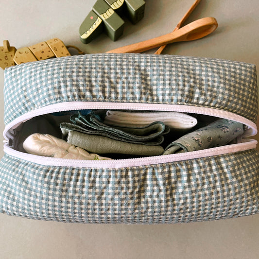 Vista desde arriba del neceser abierto donde se aprecia su amplia capacidad; muselinas de color verde con florecitas, verde liso y blanca; pañal de bebé. decoración con juguetes de madera .