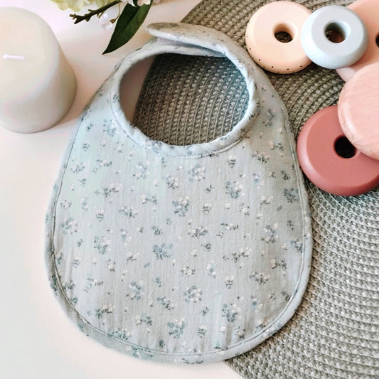 babero de delicada muselina en color verde menta combinado con flores azules; cierre en velcro adaptable al crecimiento del bebé.
