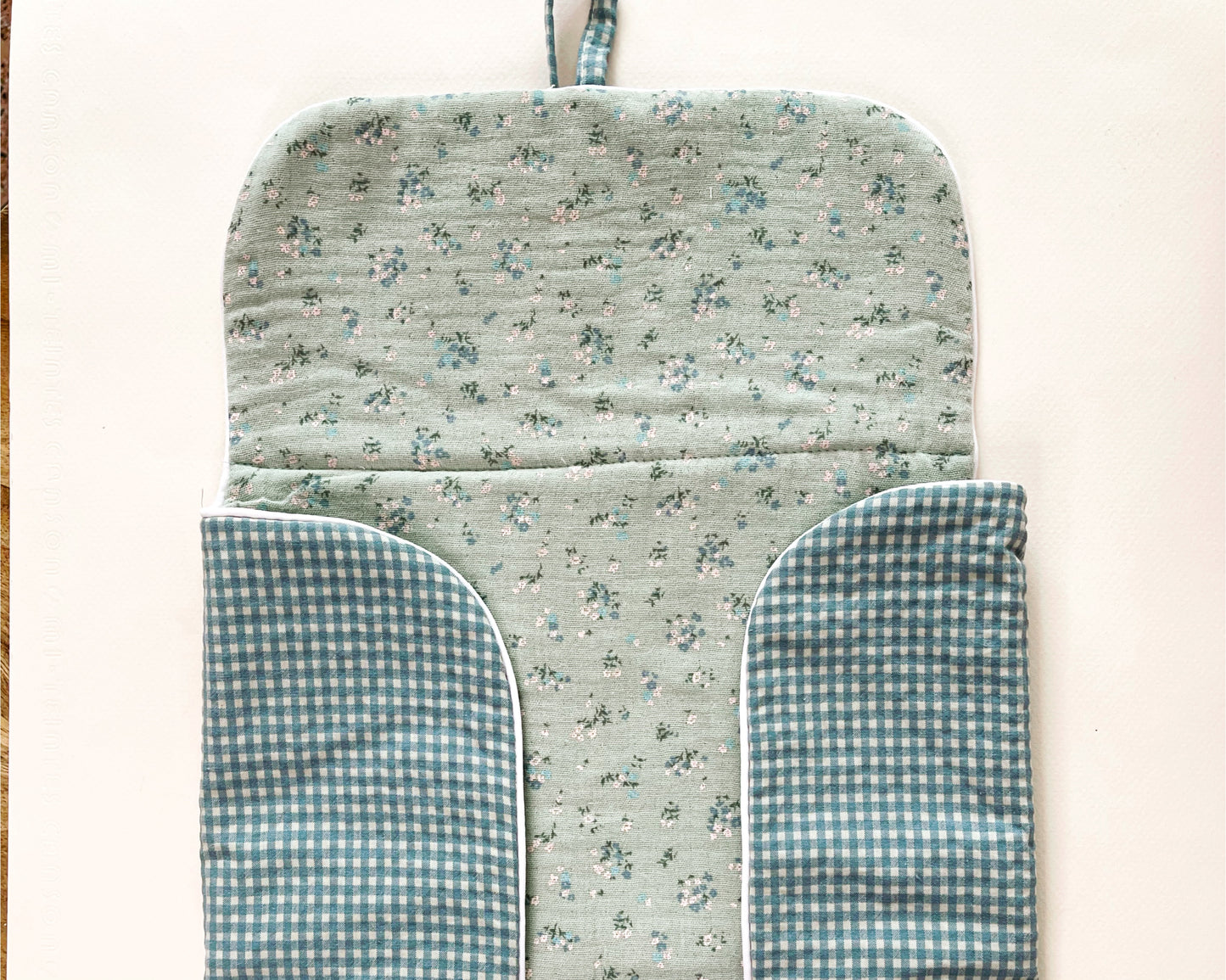 Vista del cambiador de paseo abierto donde se aprecia el interior de muselina verde con florecitas azules y el exterior de estampado cuadros en verde y blanco. Cierre con lazo.