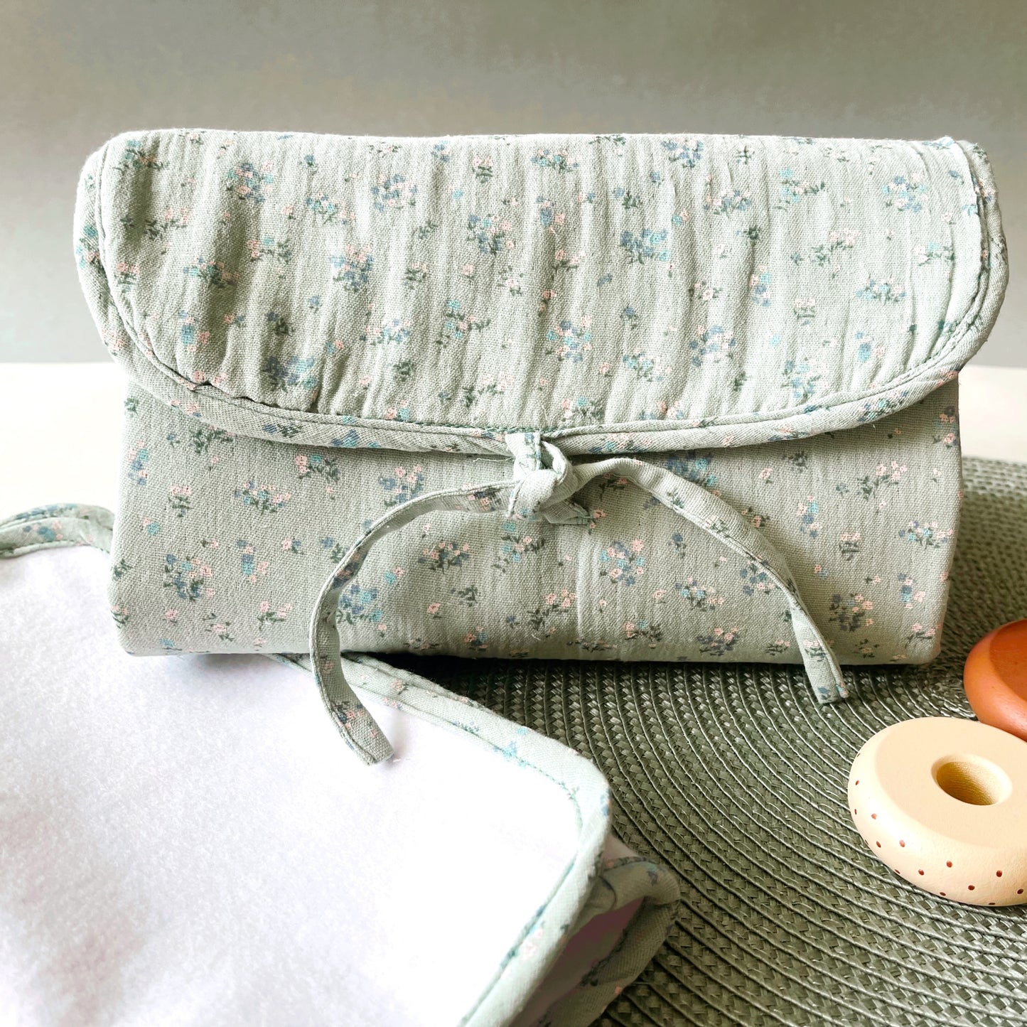 vista frontal del cambiador de paseo de muselina en color verde menta con flores azules y blancas: cierre con lazo; alfombra para cambiar al bebé de color blanco y de suave guata impermeable 