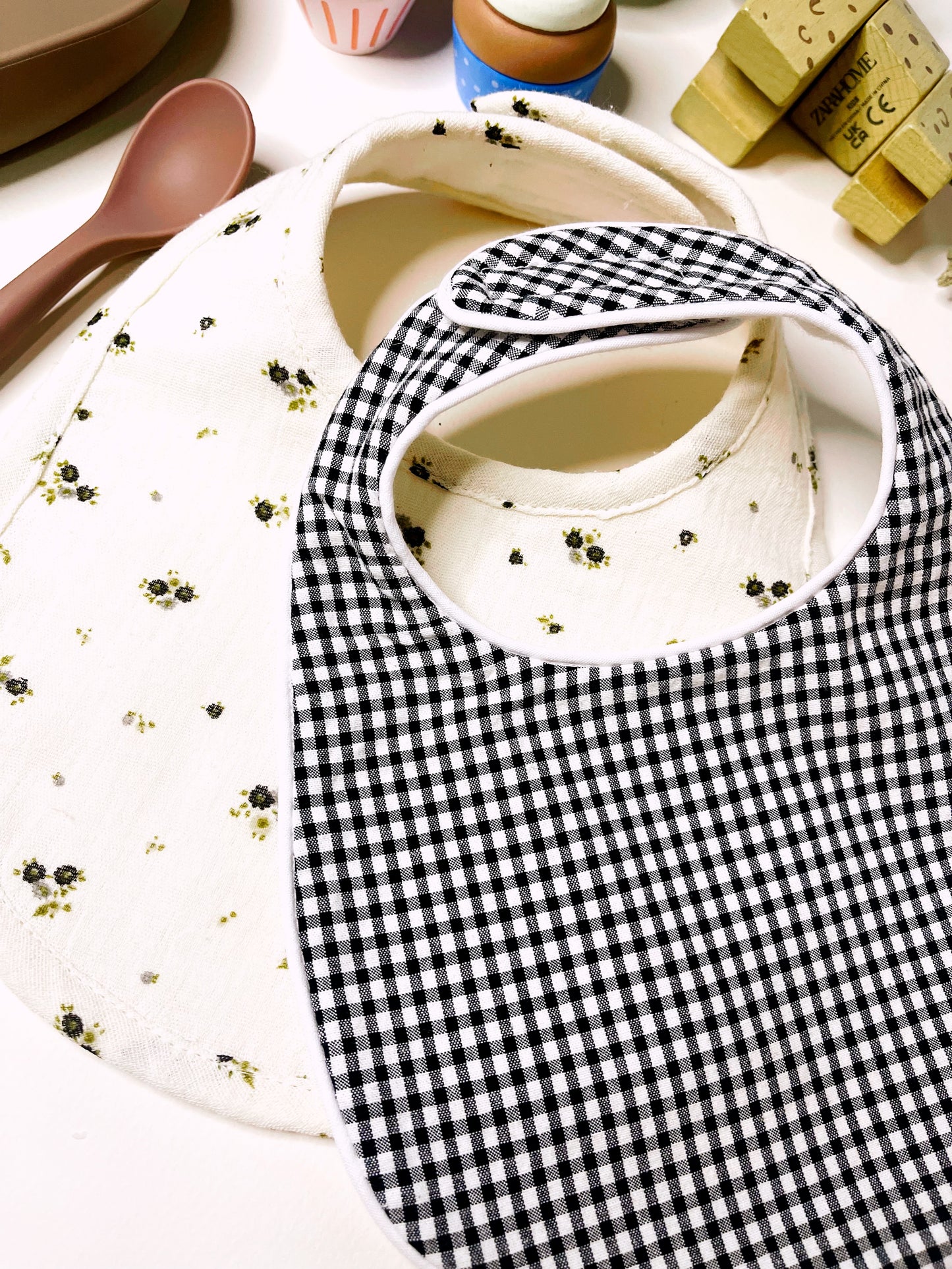 Vista de cerca del conjunto de babero en estampado de cuadros negro y blanco junto con otro babero de muselina beis con flores grises y verdes. Decoración de juguetes de madera.