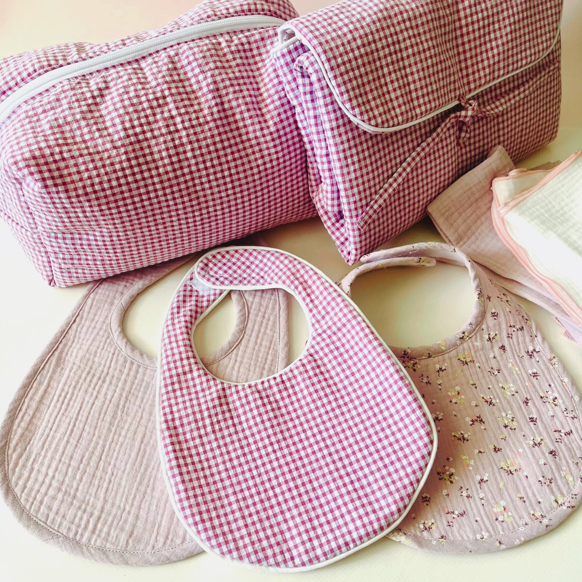 Conjunto de tres baberos, el del centro de estampado en cuadros rosa y blanco, a la izquierda en muselina malva liso y a la derecha en muselina malva con florecitas; detrás se sitúan el neceser y el cambiador de paseo, ambos en cuadros rosa y blanco.