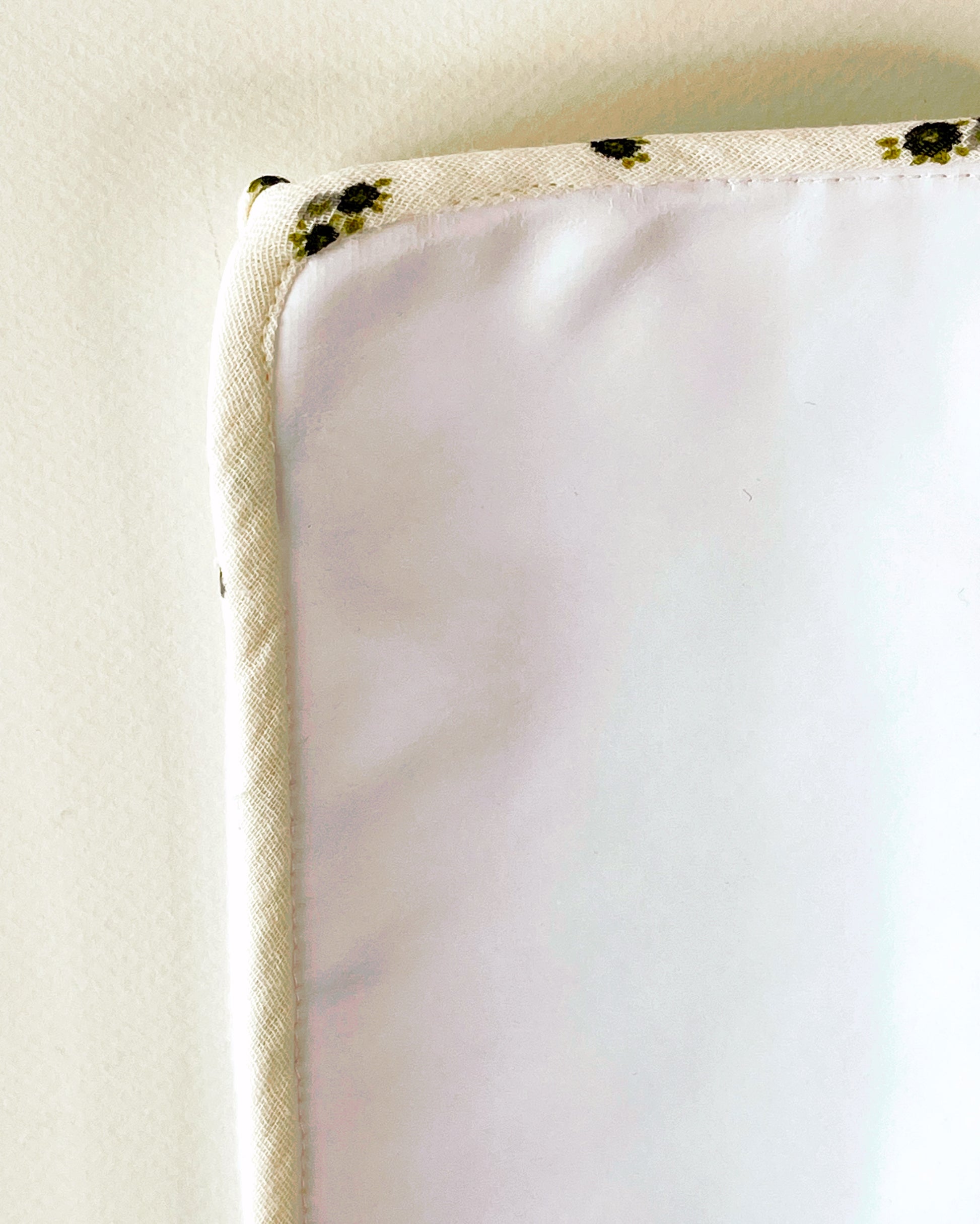 Parte trasera de la alfombra para cambiar al bebé de suave guata impermeable; acabado en cordón de vibo de muselina beis con florecitas grises y verdes.