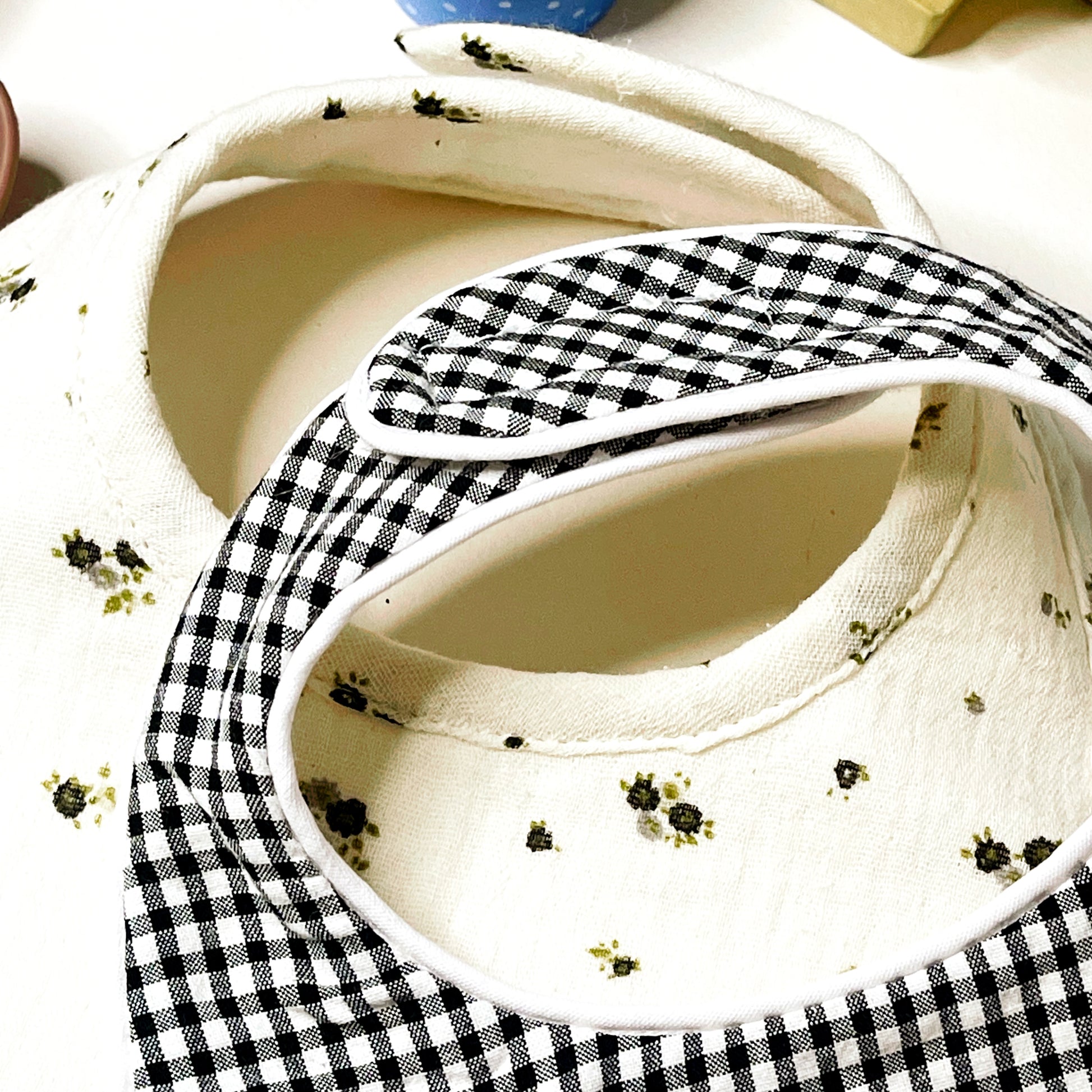 Detalle muy de cerca de los baberos en estampado de cuadros blanco y negro y del babero muselina beis con flores grises y verdes: cierre con velcro y fino acabado en cordón vibo blanco