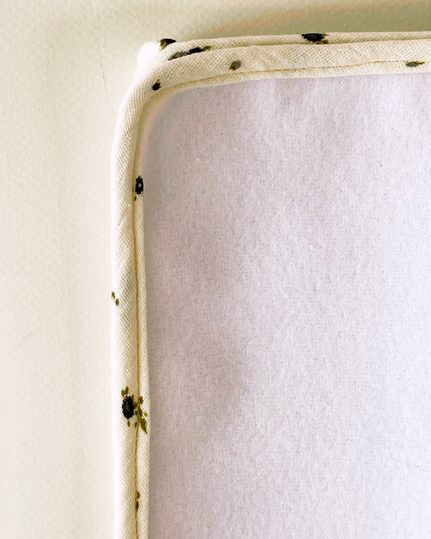 Parte delantera de la alfombra para cambiar al bebé de suave guata. Acabado en cordón de vibo de muselina beis con flores de color gris y verde.