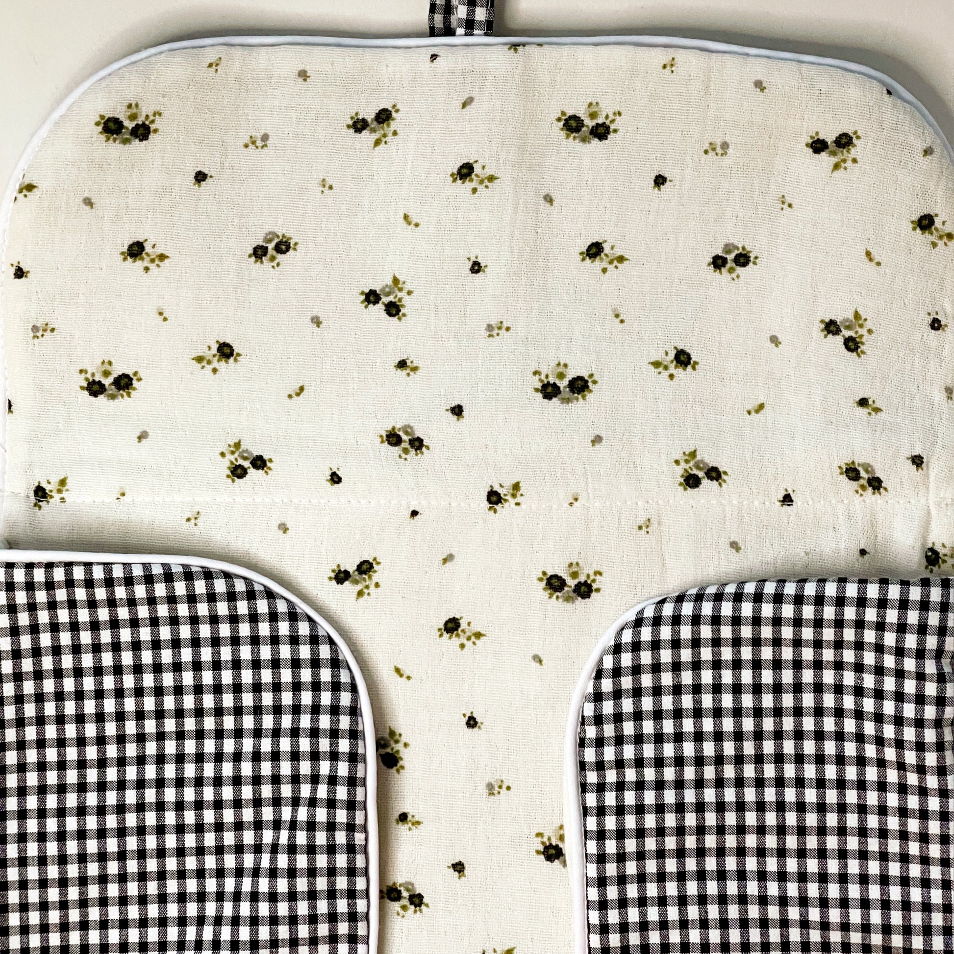 Lado interno del cambiador de paseo en muselina de color beis con flores grises y verde y en la parte exterior estampado de cuadros en color negro y blanco