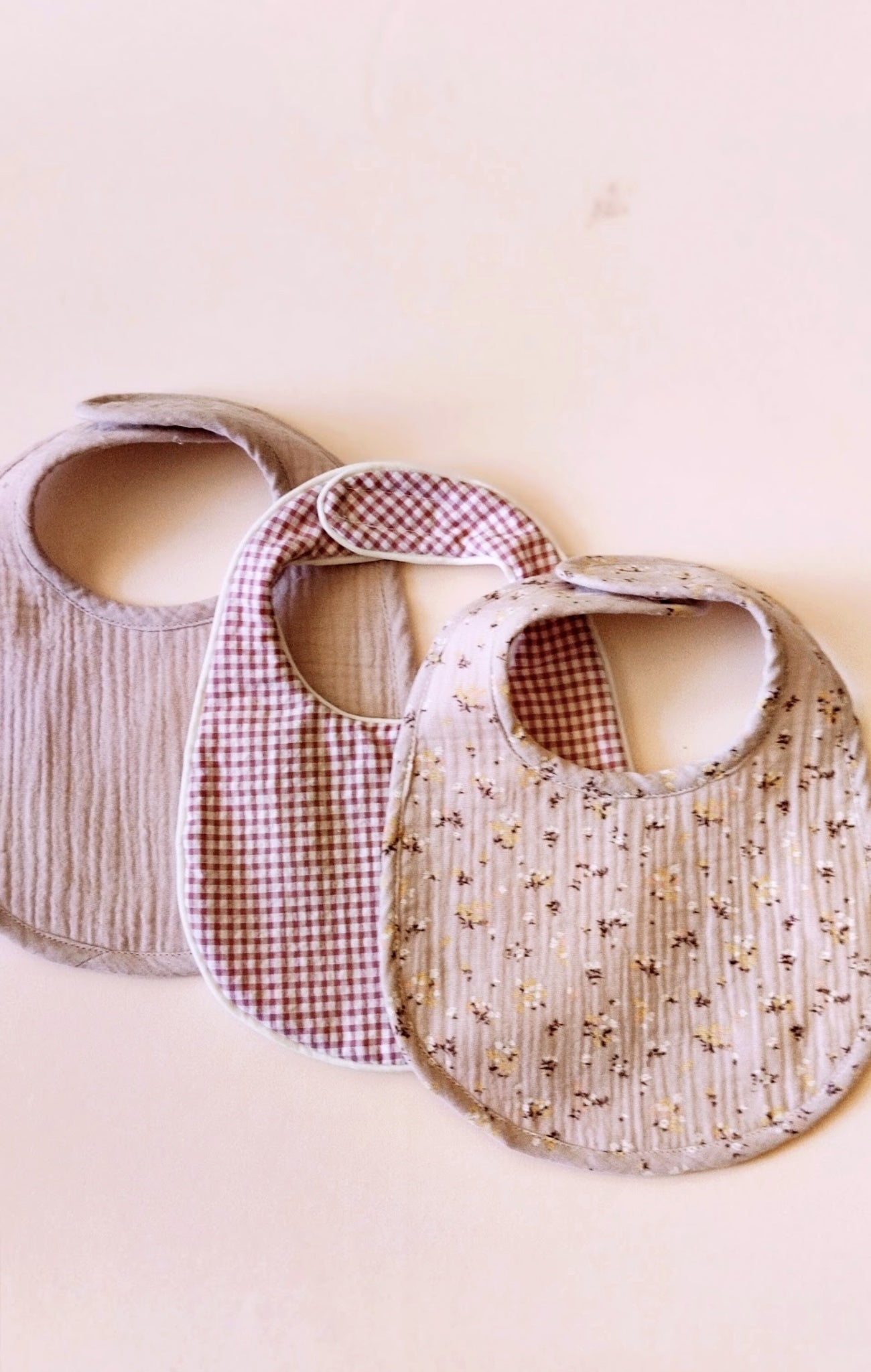 Conjunto de tres baberos de muselina, el de la izquierda en color malva liso, el del centro en estampado de cuadros rosa y blanco y el de la derecha de muselina malva con flores amarillas y burdeos 