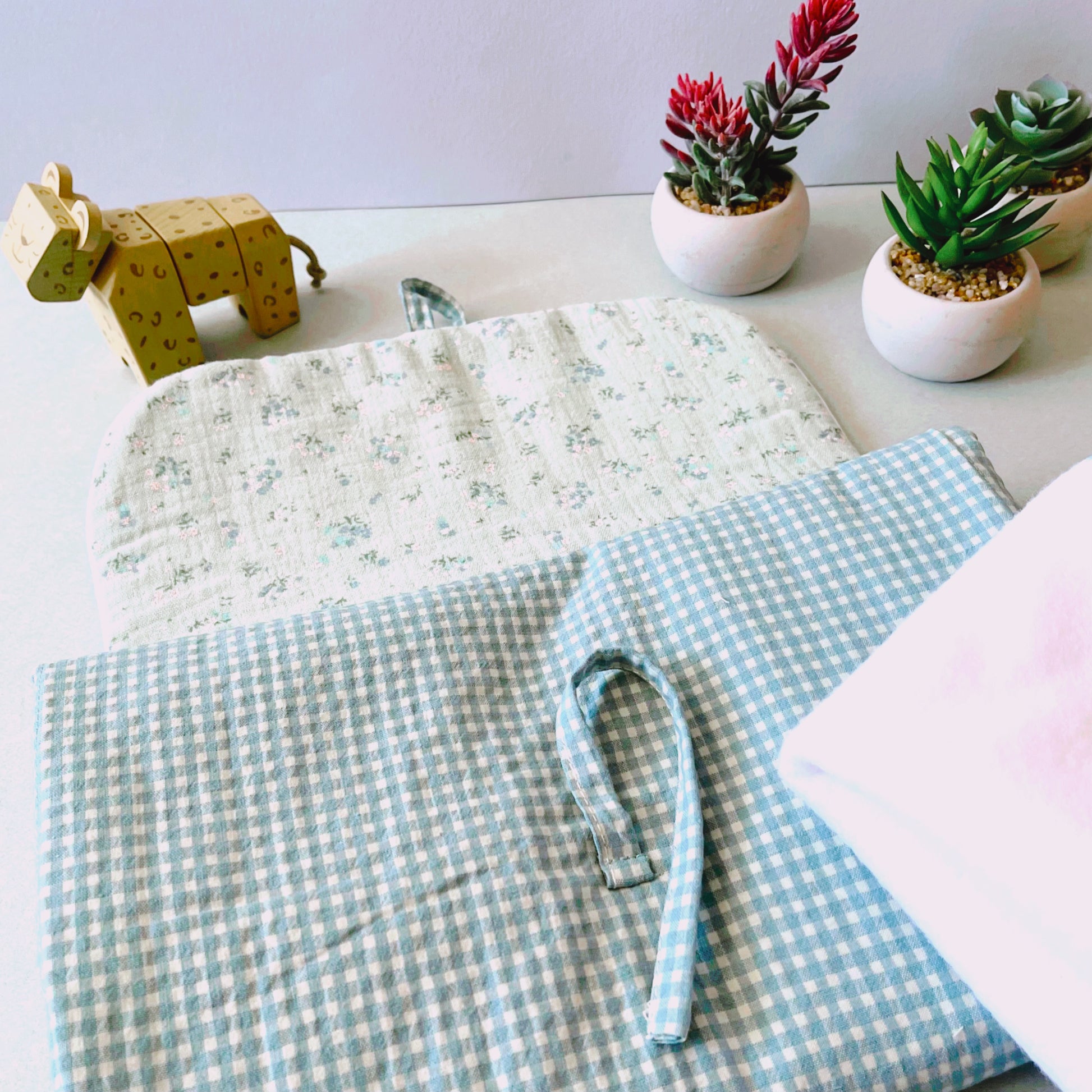 Cambiador abierto donde se aprecia la parte interior de suave muselina de color verde con florecitas; cierre con lazos y muselina de color blanco liso. Decorado con juguete de madera y plantas