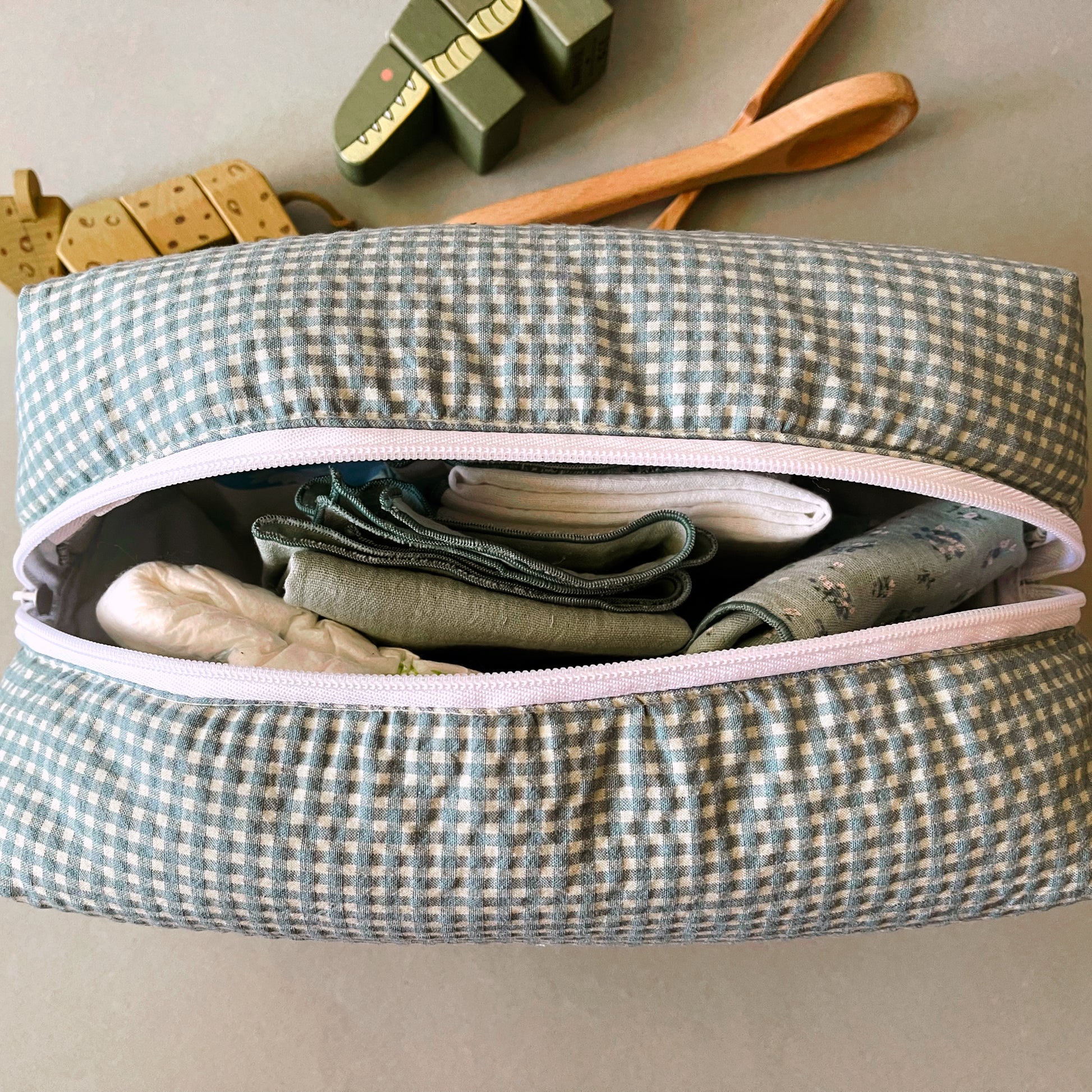 Vista desde arriba del neceser abierto donde se aprecia su amplia capacidad; muselinas de color verde con florecitas, verde liso y blanca; pañal de bebé. decoración con juguetes de madera .