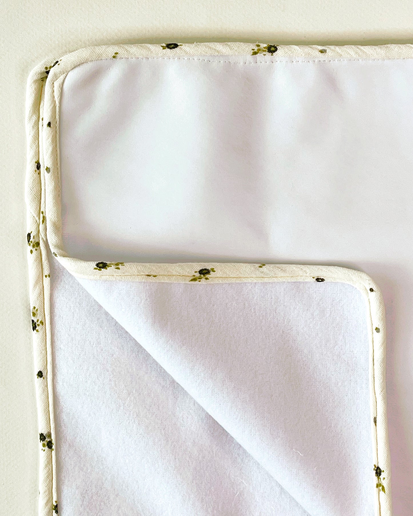 Vista de la alfombrilla que acompaña al cambiador donde se aprecia la guata, suave y delicada para apoyar al bebé, y la guata impermeable para evitar que se pueda calar el cambiador. Los bordes están perfectamente acabados en una muselina beis con flores igual que el cambiador