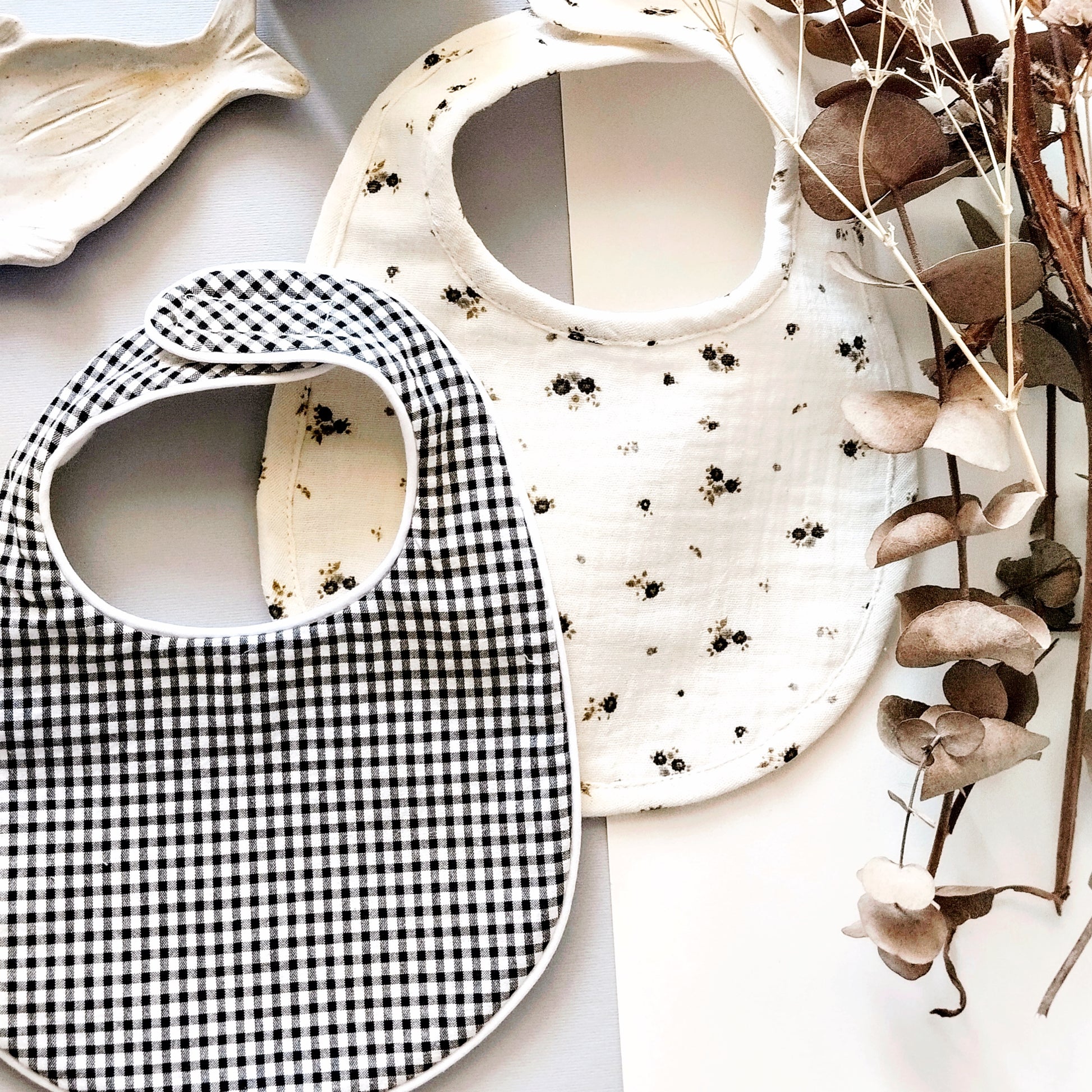 Conjunto de babero en estampado de cuadros negro y blanco junto con otro babero de muselina beis con flores grises y verdes. Decoración de flores