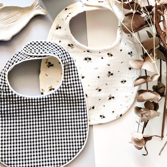 Conjunto de babero en estampado de cuadros negro y blanco junto con otro babero de muselina beis con flores grises y verdes. Decoración de flores