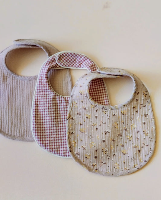 conjunto d elos tres baberos que componen la colección PEC, uno de muselina color malva con florecitas, el otro en estampado Vichy de color rosa y blanco y el tercero de color malva liso