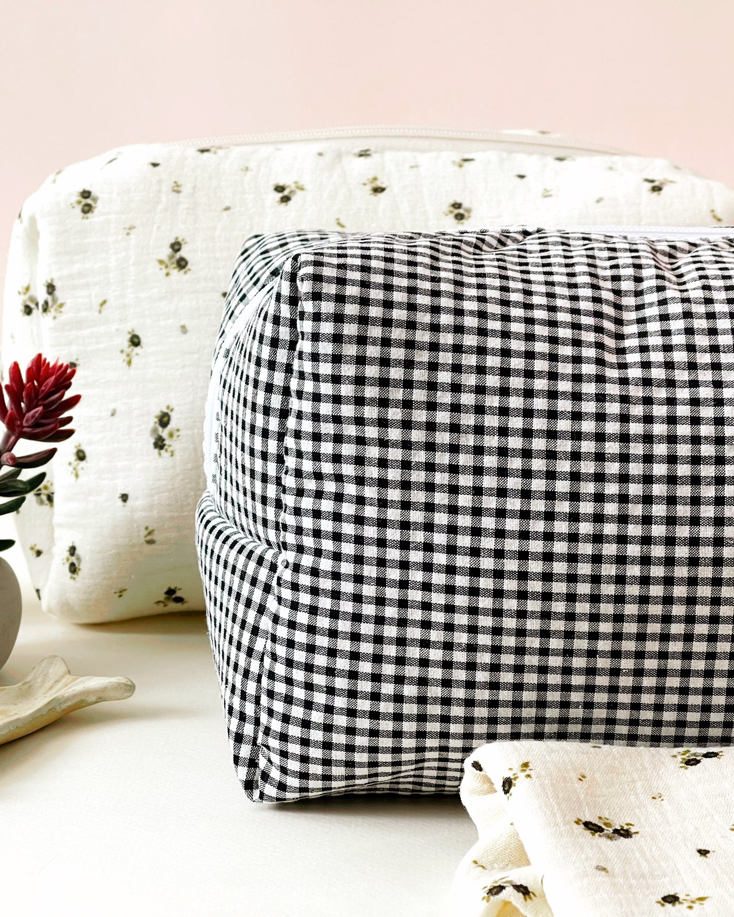 Vista de frente del neceser de estampado de cuadros blanco y negro; detrás está situado un neceser de muselina beis con flores grises y verdes ; muselina de color beis con florecitas grises y verdes.