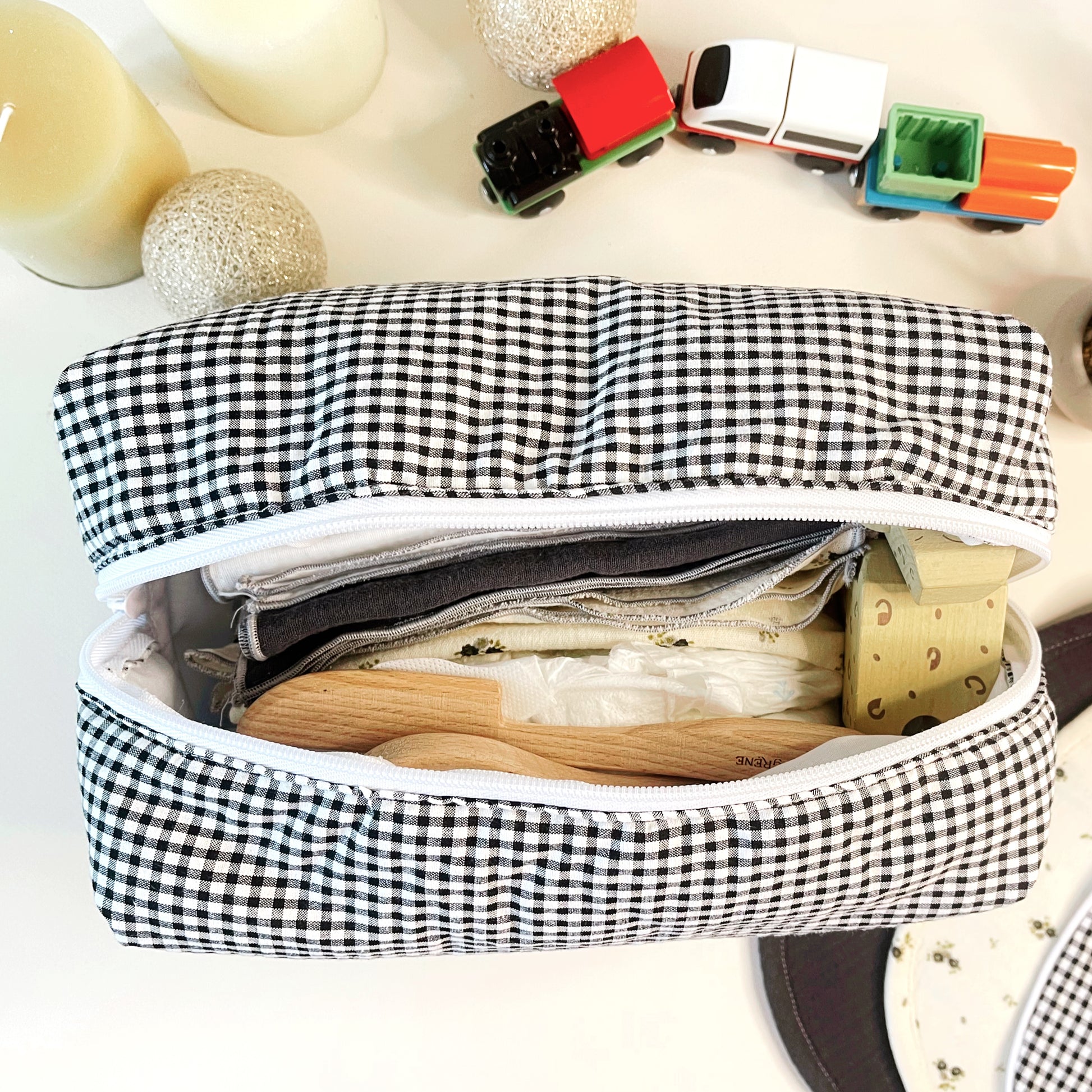 Vista desde arriba del neceser abierto y en su interior hay muselinas de color gris liso y blanco liso, juguetes de madera. Amplia apertura de la cremallera para facilitar su uso.