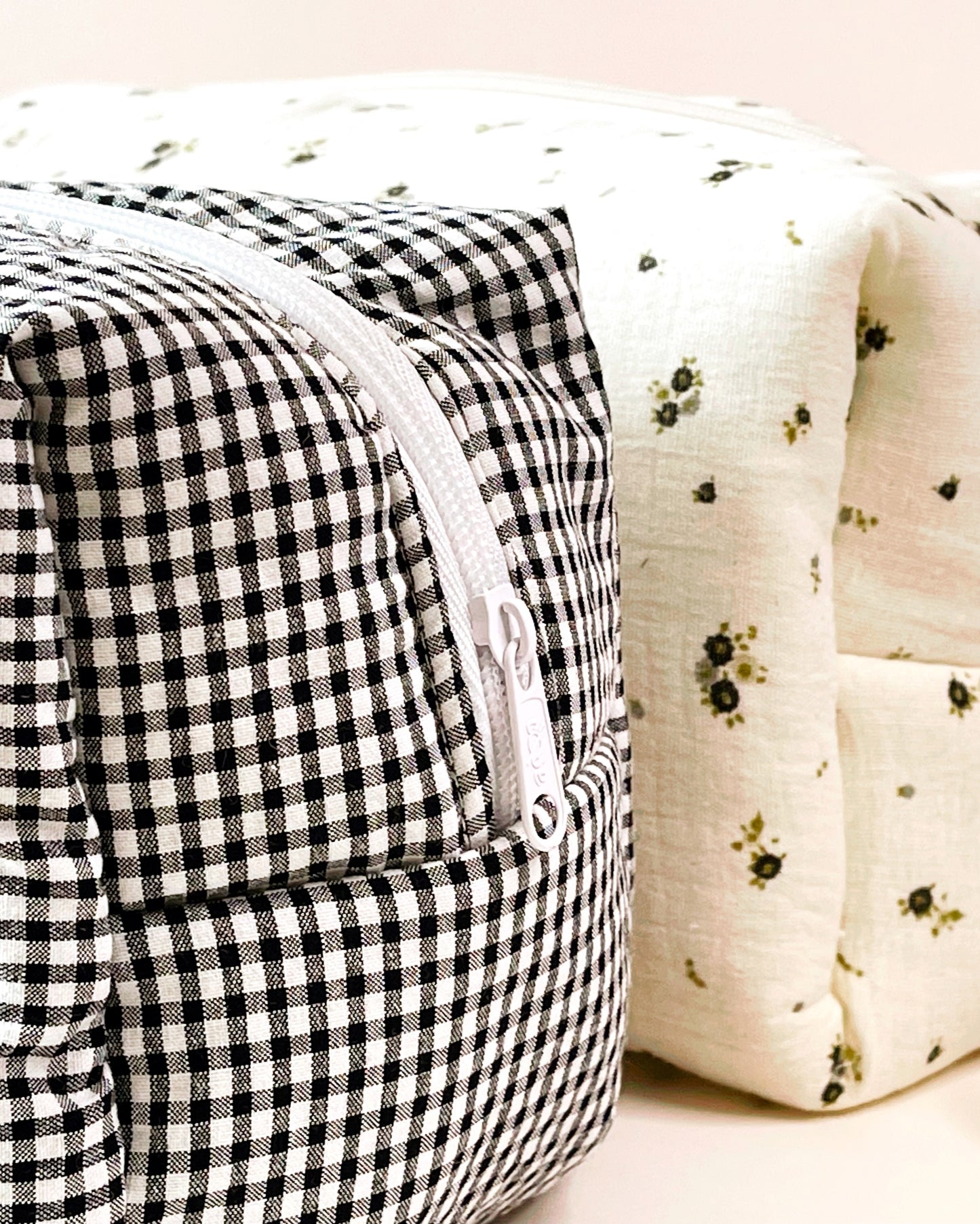 Parte lateral del neceser de estampado de cuadros blanco y negro con cierre en cremallera de color blanca; al lado está situado un neceser de muselina beis con flores grises y verdes.