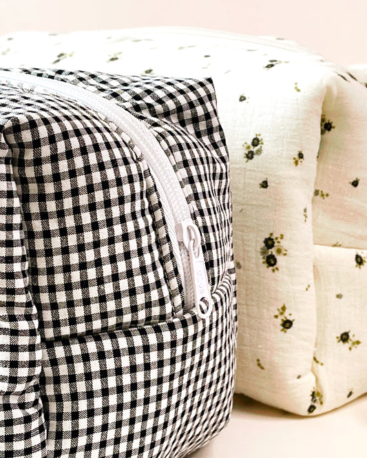 Parte lateral del neceser de estampado de cuadros blanco y negro con cierre en cremallera de color blanca; al lado está situado un neceser de muselina beis con flores grises y verdes.