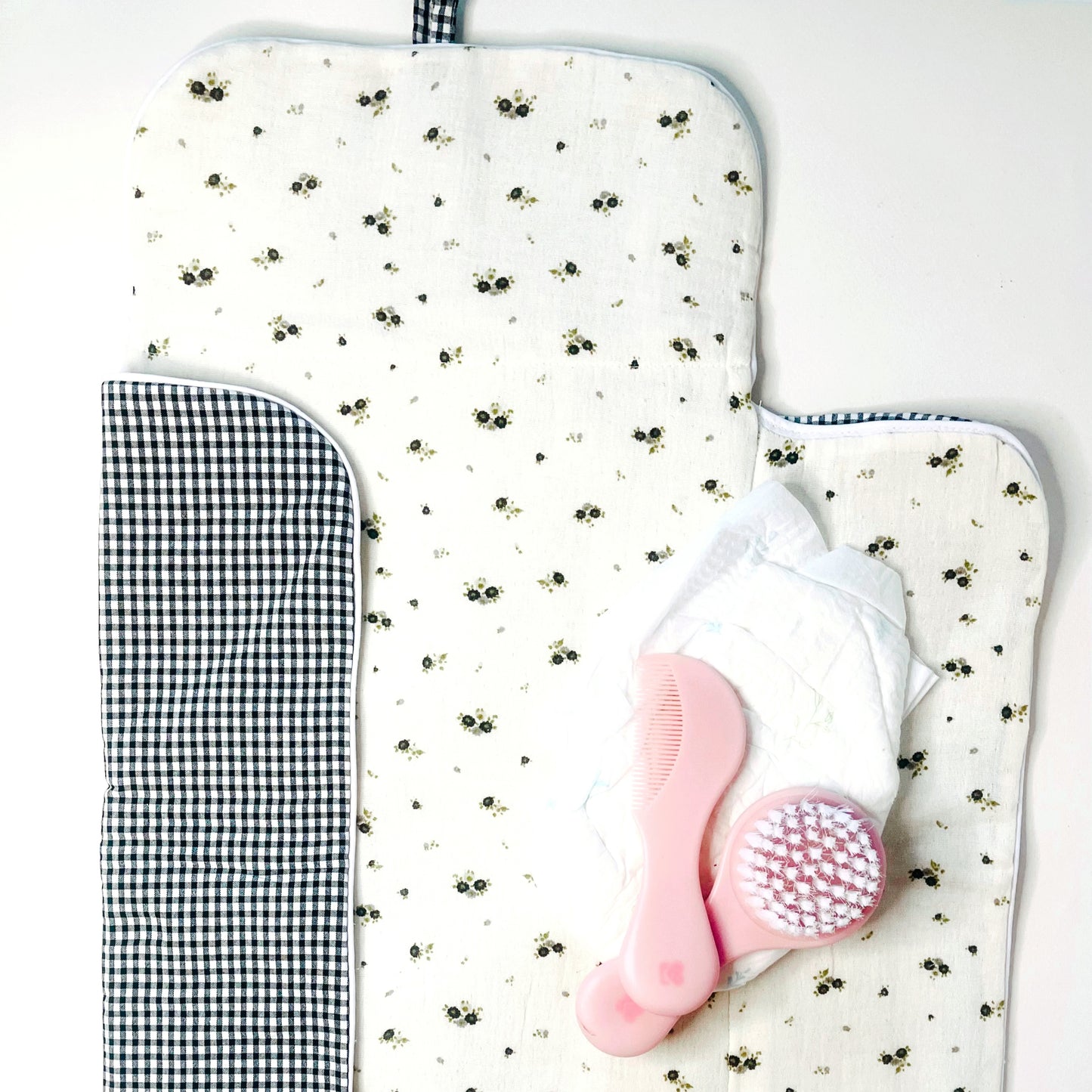 Cambiador de paseo abierto; en el interior tejido  en muselina de color beis con flores grises y verde y en la parte exterior estampado de cuadros en color negro y blanco. decoración pañales y peine de bebé