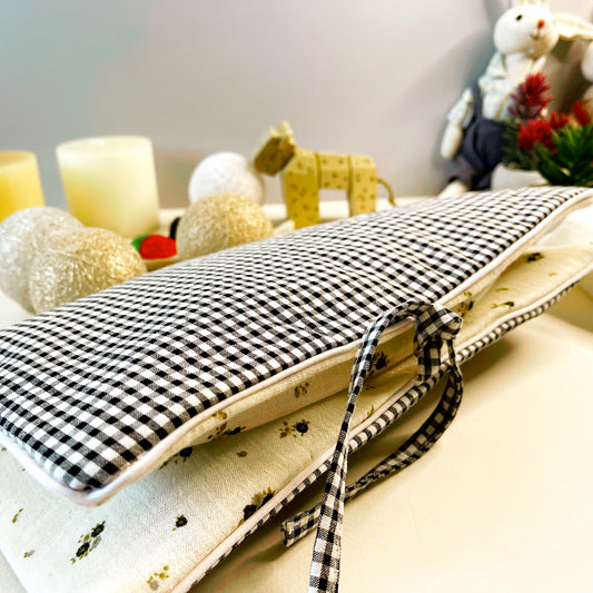 Vista lateral del porta toallitas cerrado donde se aprecia la combinación de teñlas; en la parte exterior estampado de cuadros blanco y negro y en la parte interior muselina beis con flores grises y verdes; acabado en un cordón en vibo blanco y cierre con dos lazos