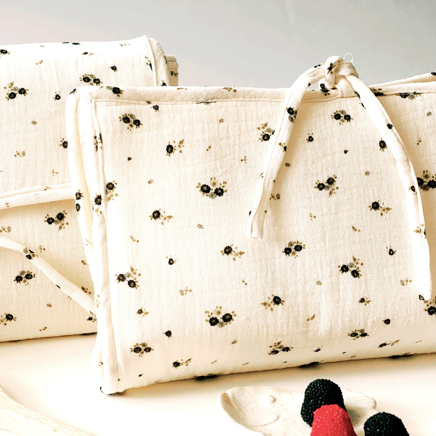 Vista de frente del porta toallitas en muselina de color beis con florecitas gris y verde; cierre con lazo. Detrás, está el cambiador del mismo tejido