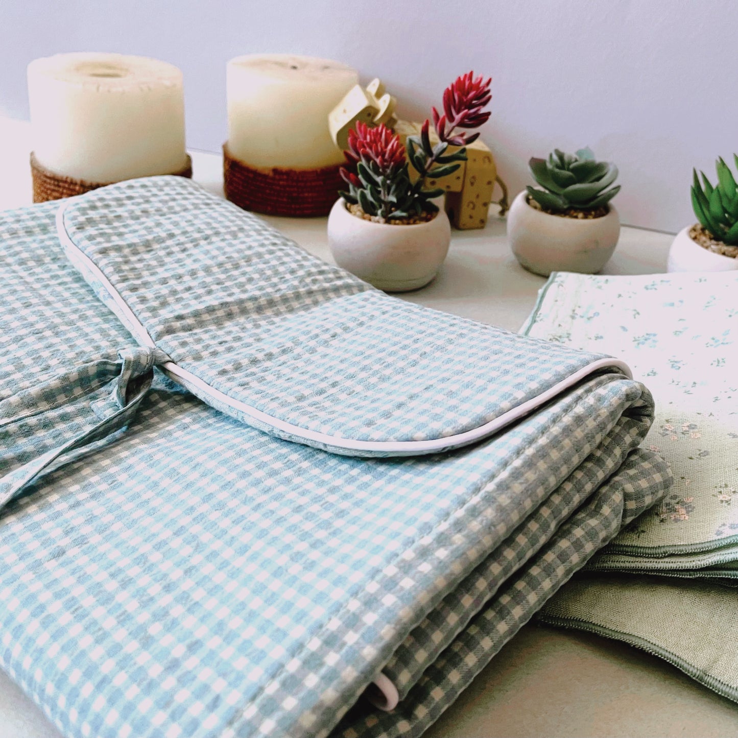 Cambiador de paseo plegado de estampado en color verde menta y blanco, cierre con dos lazos y muselina de color verde con florecitas azules y muselina verde liso. Composición de juguetes y plantitas